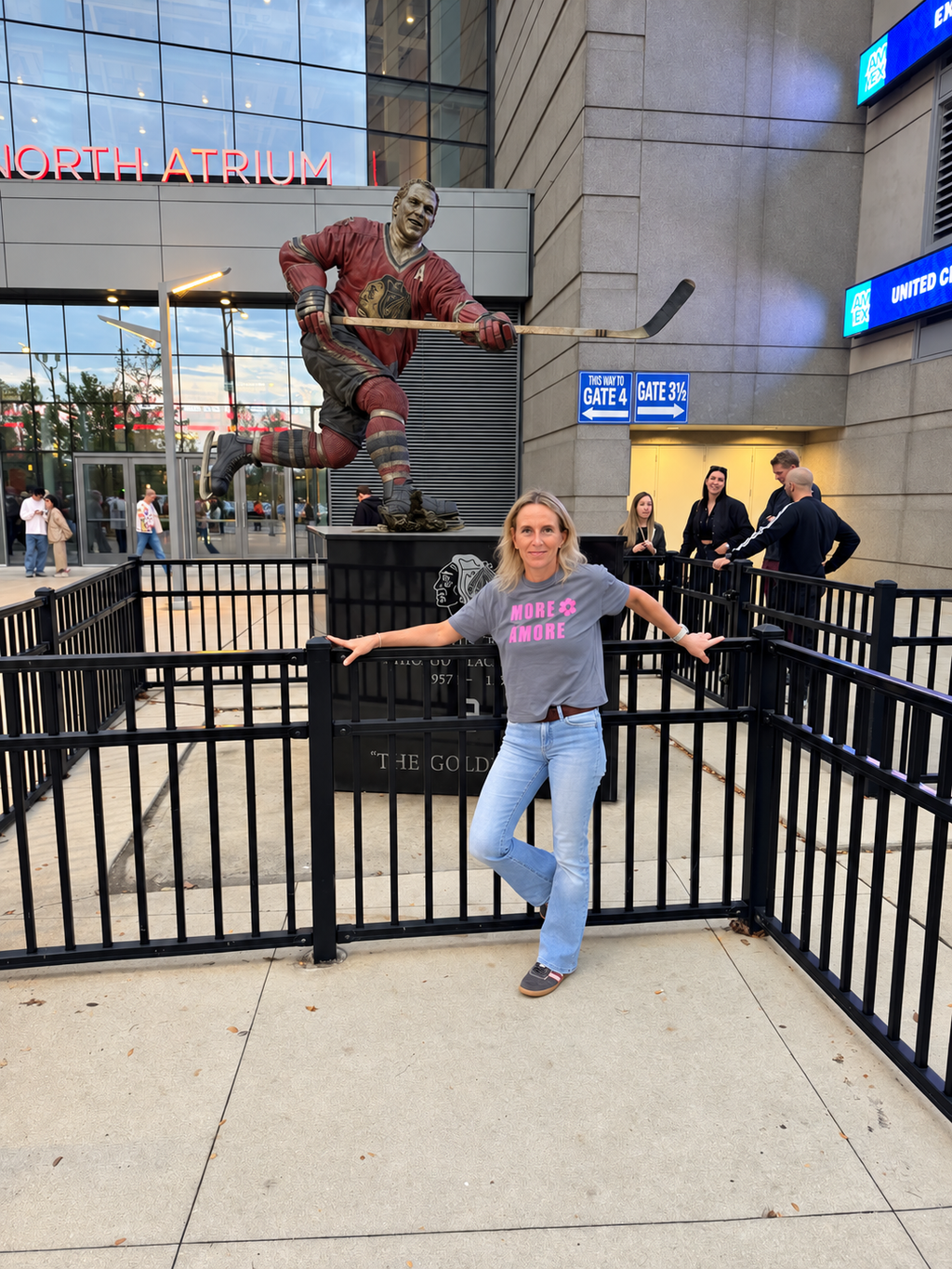 Karin trägt das MORE AMORE Vintage Blue Cropped T-Shirt mit pinkem Frontprint vor einer Eishockey-Statue in Chicago.