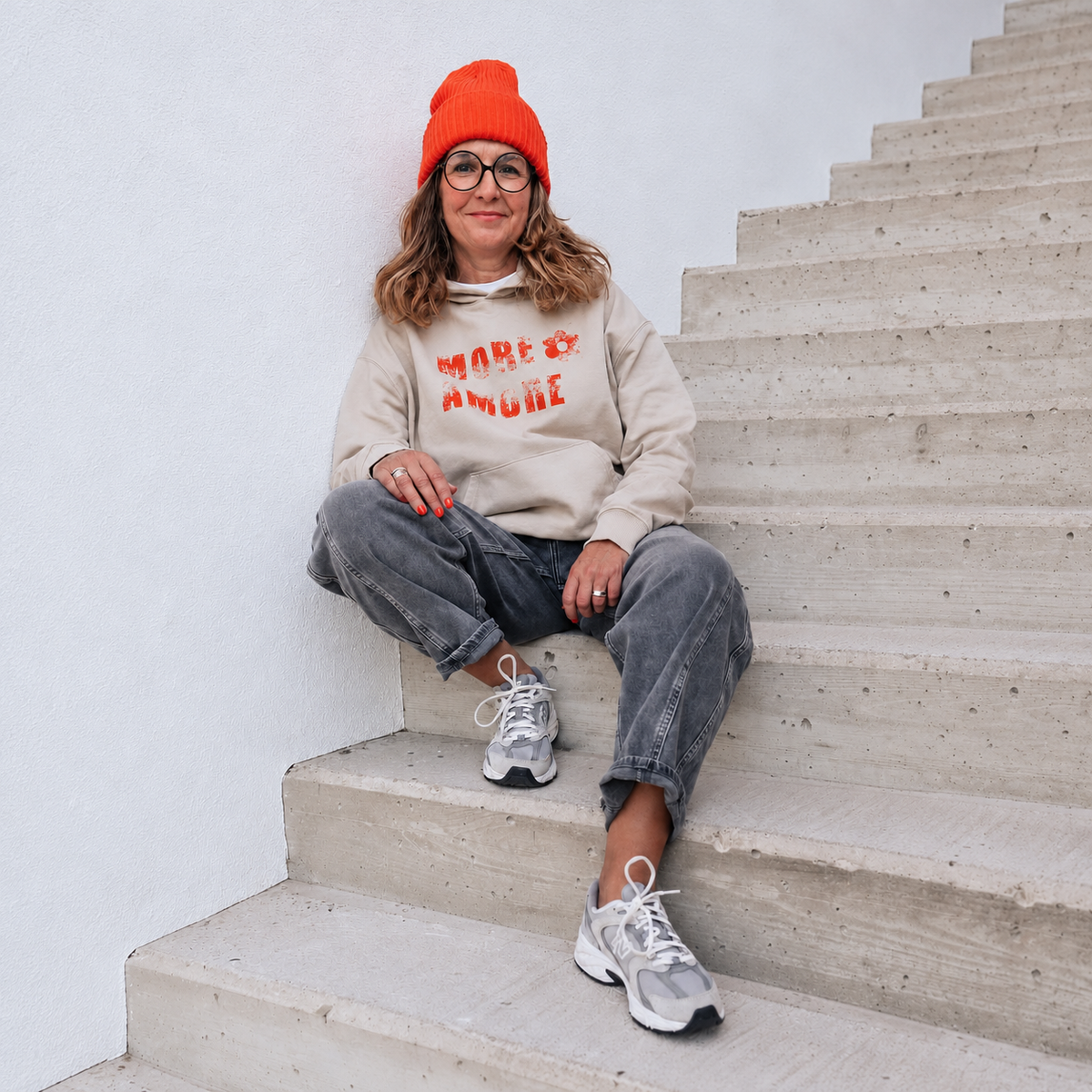 Marion sitzt lächelnd auf einer Betontreppe und trägt den MORE AMORE Boxy Unisex-Hoodie in Warm Sand mit rotem Floral-Print.